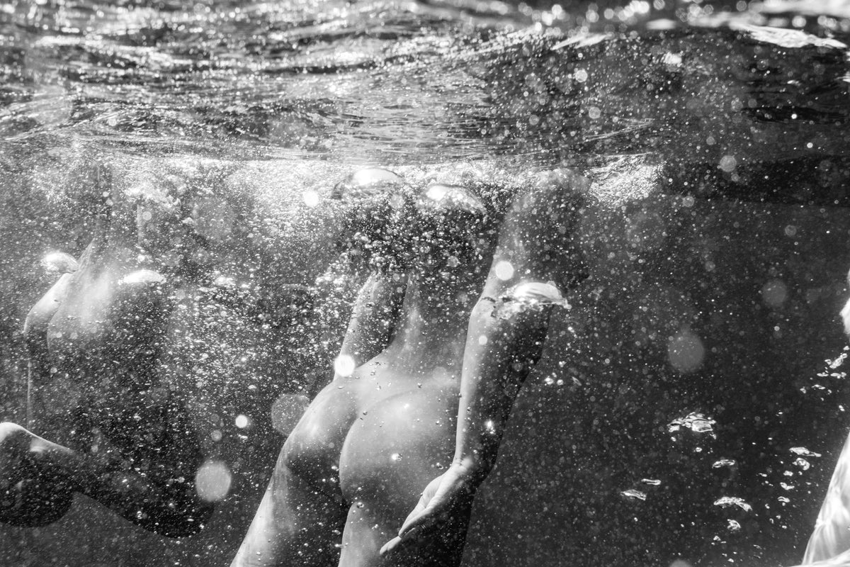 A figure rises toward the water's surface in a swirl of bubbles, light refractions and movement, other figures visible beyond. A striking underwater fine art print by Emma Maggs.