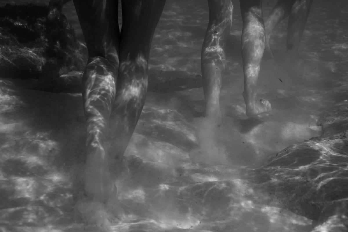 Sirens Call – black and white underwater fine art photograph looking up at the legs and feet of multiple female figures standing in shallow water, light refracting across the sandy floor, by Emma Maggs